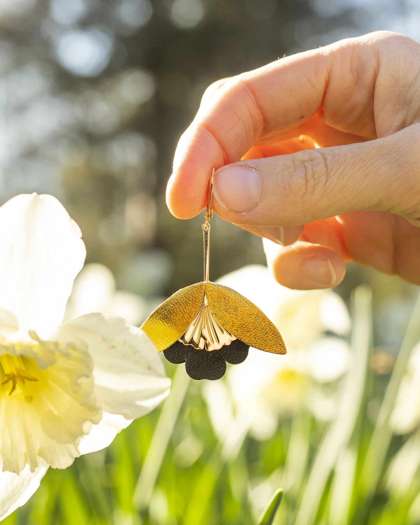 Boucles d'oreilles Fleurs de Ginkgo cuir doré et noir tenues à la main en extérieur avec fleurs en arrière-plan