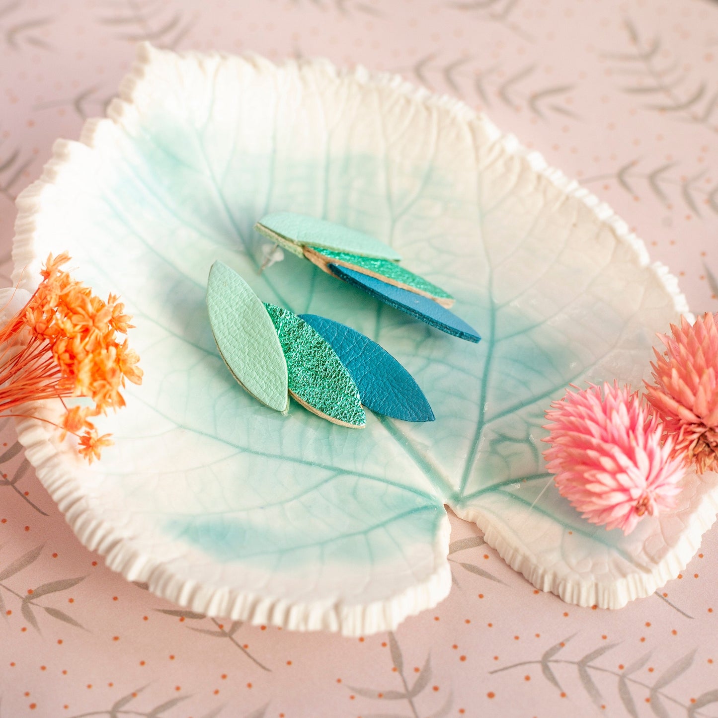Boucles d'oreilles cuir vert bleu en forme de feuilles posées sur une assiette feuille avec fleurs décoratives