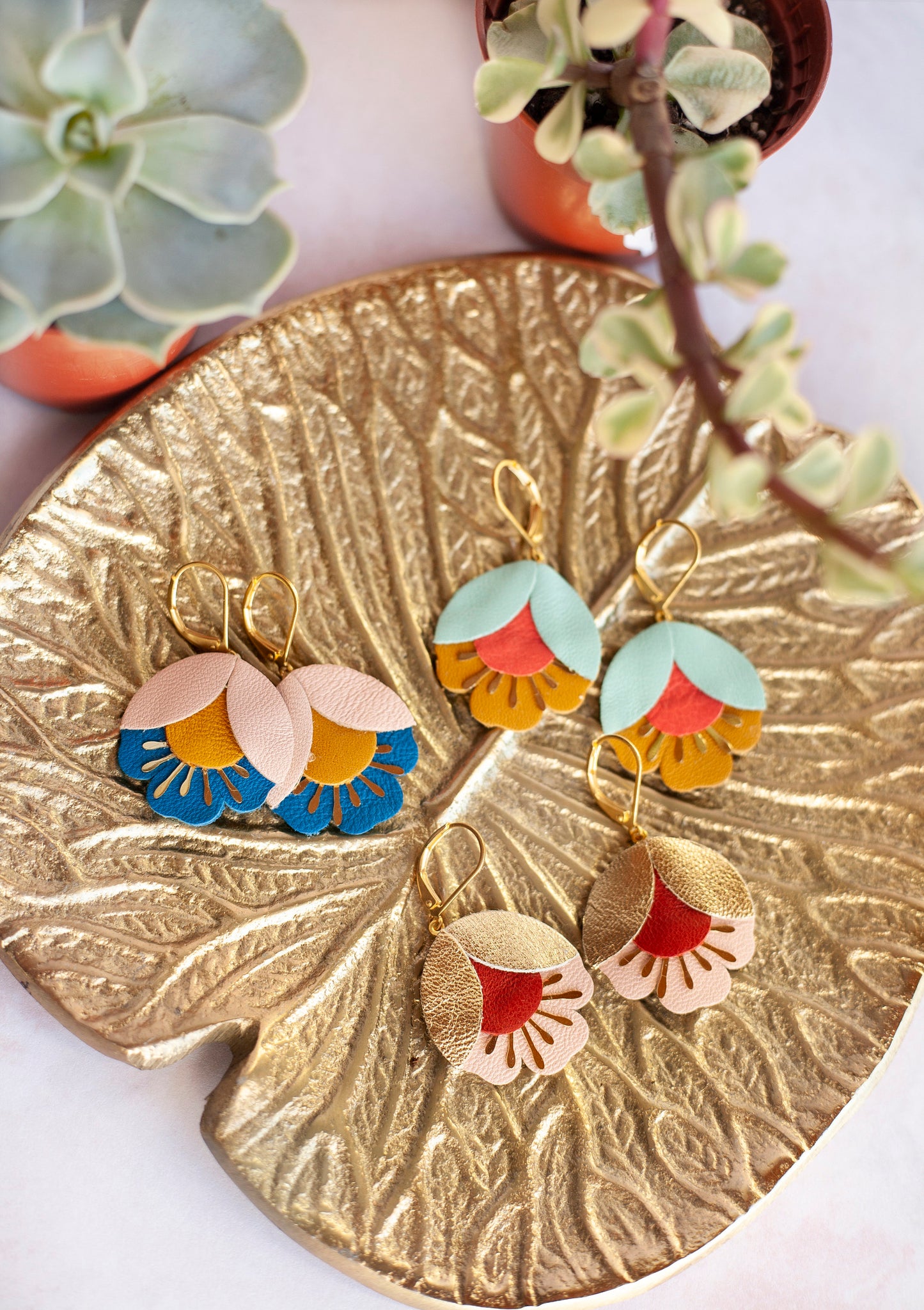 Boucles d'oreilles fleurs de cerisier cuir doré rouge et rose présentées sur un plateau doré avec des plantes succulentes