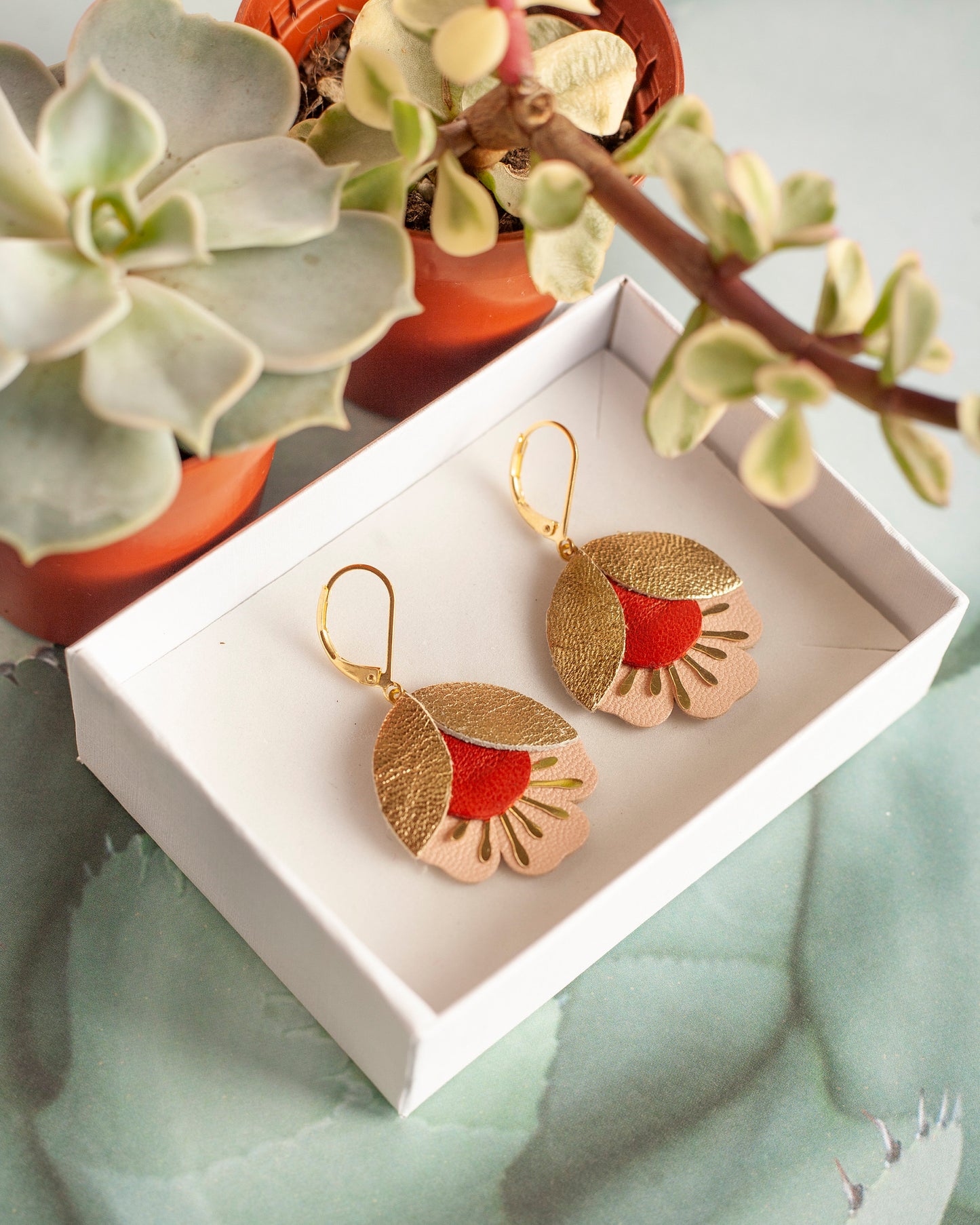 Boucles d'oreilles fleurs de cerisier cuir doré rouge et rose présentées dans une boîte blanche avec décor de plantes succulentes