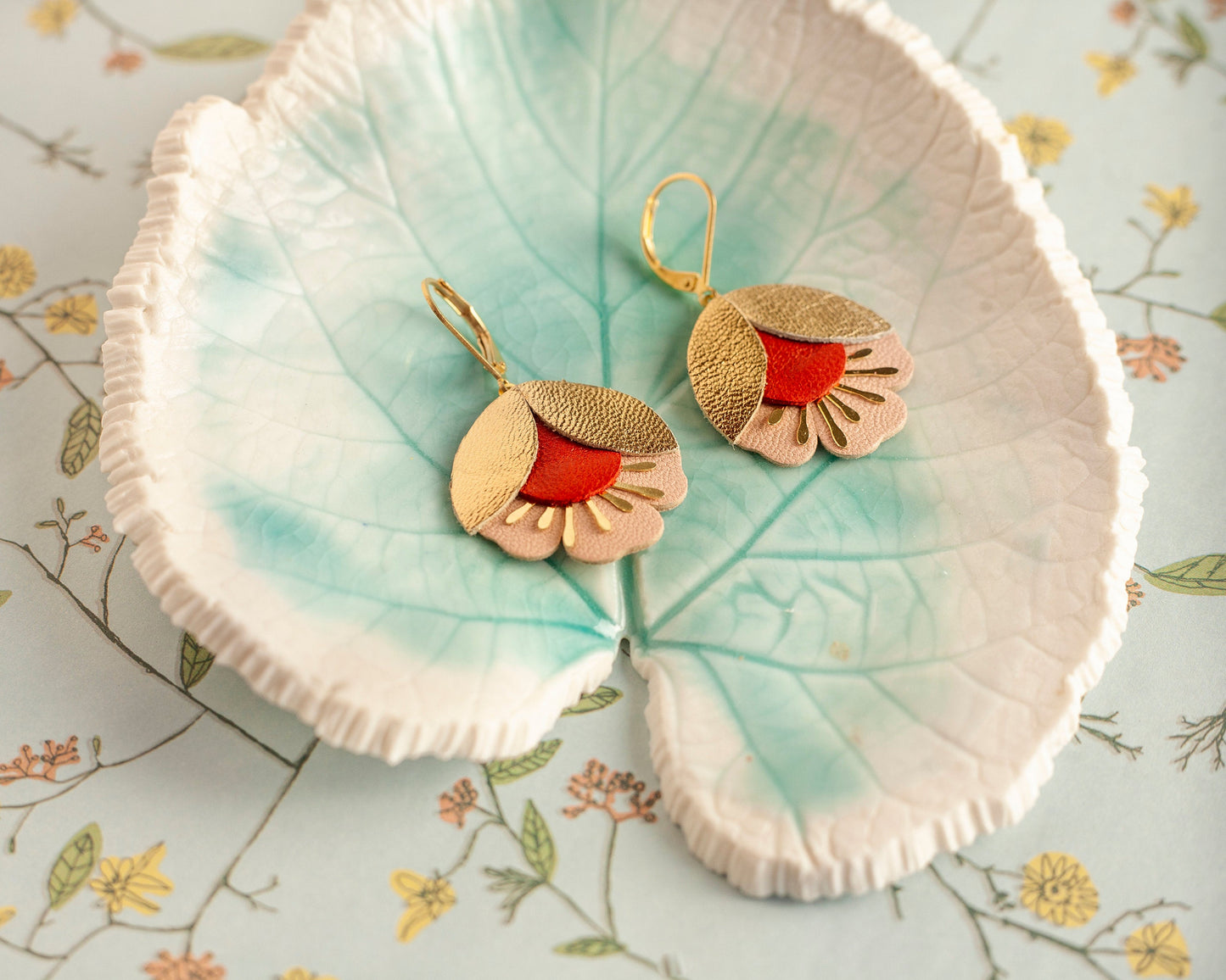 Boucles d'oreilles fleurs de cerisier cuir doré rouge et rose posées sur un plat feuille vert pâle