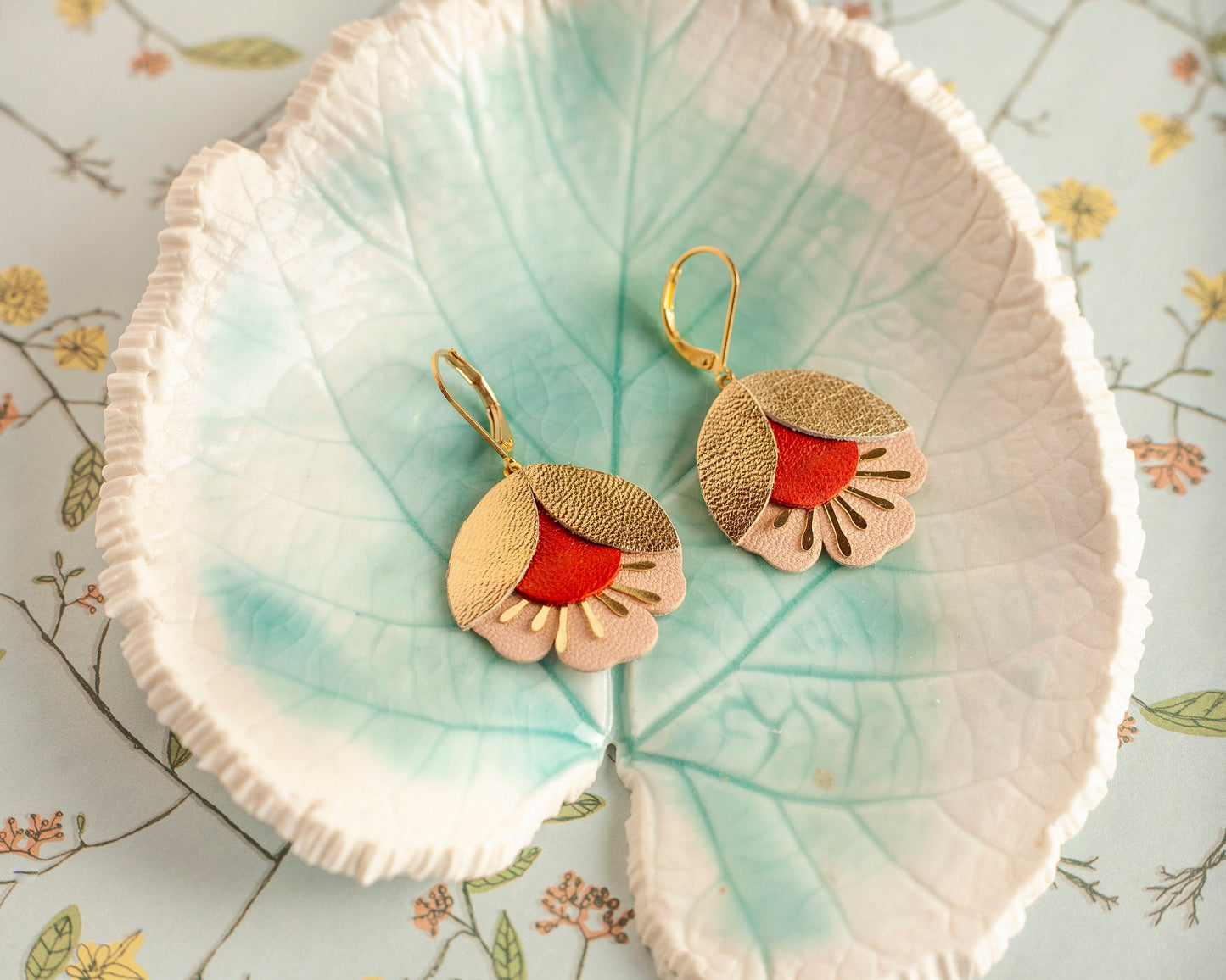 Boucles d'oreilles fleurs de cerisier cuir doré rouge et rose posées sur une coupelle en forme de feuille