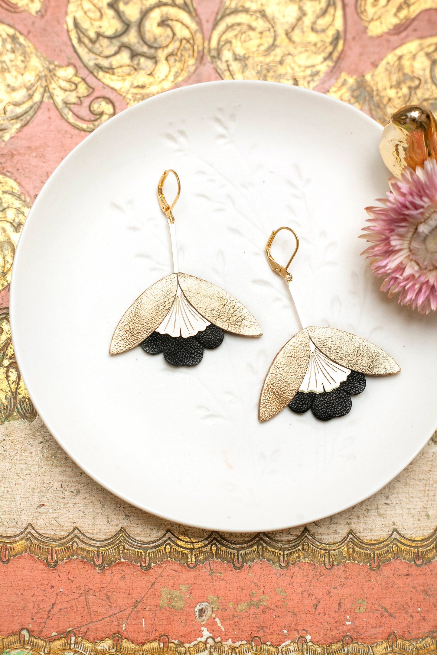 Boucles d'oreilles Fleurs de Ginkgo cuir doré et noir élégantes posées sur une assiette blanche