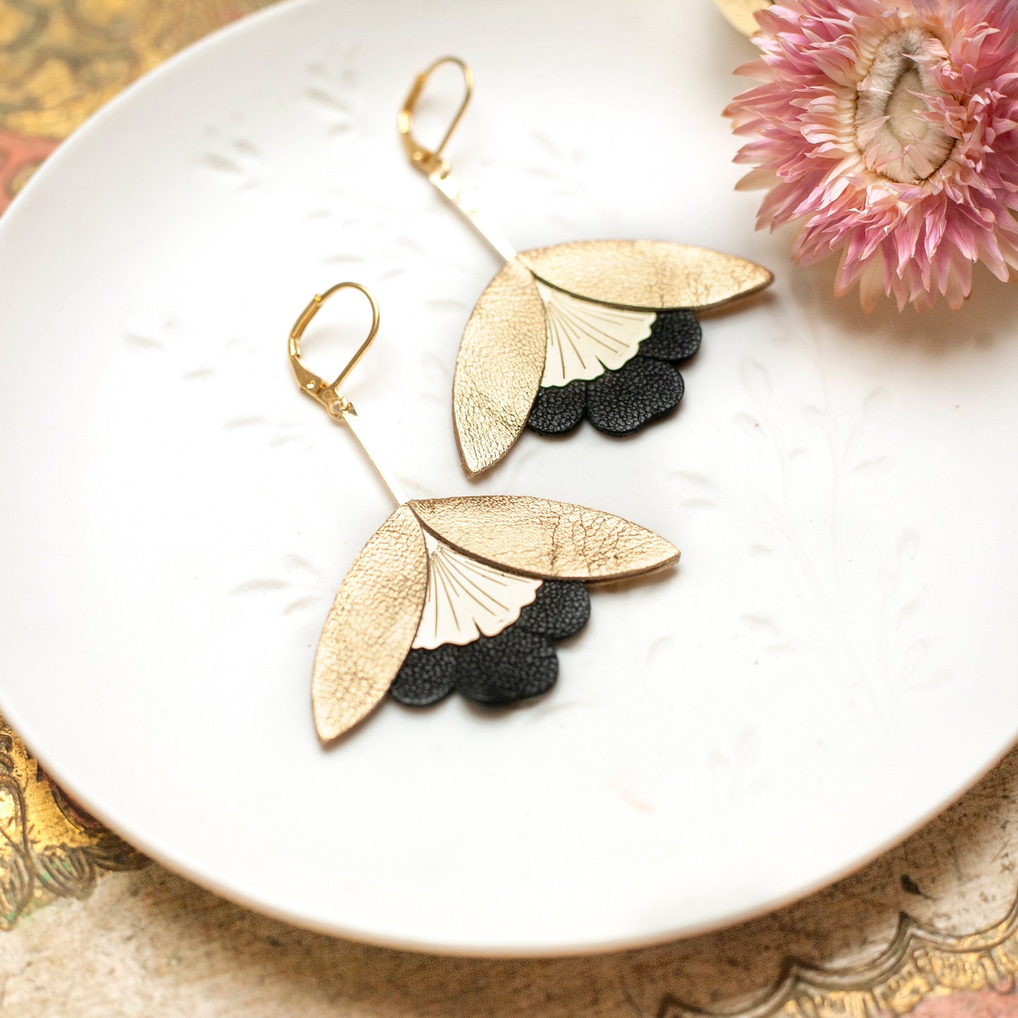 Boucles d'oreilles Fleurs de Ginkgo cuir doré et noir élégantes posées sur une assiette blanche avec une fleur rose