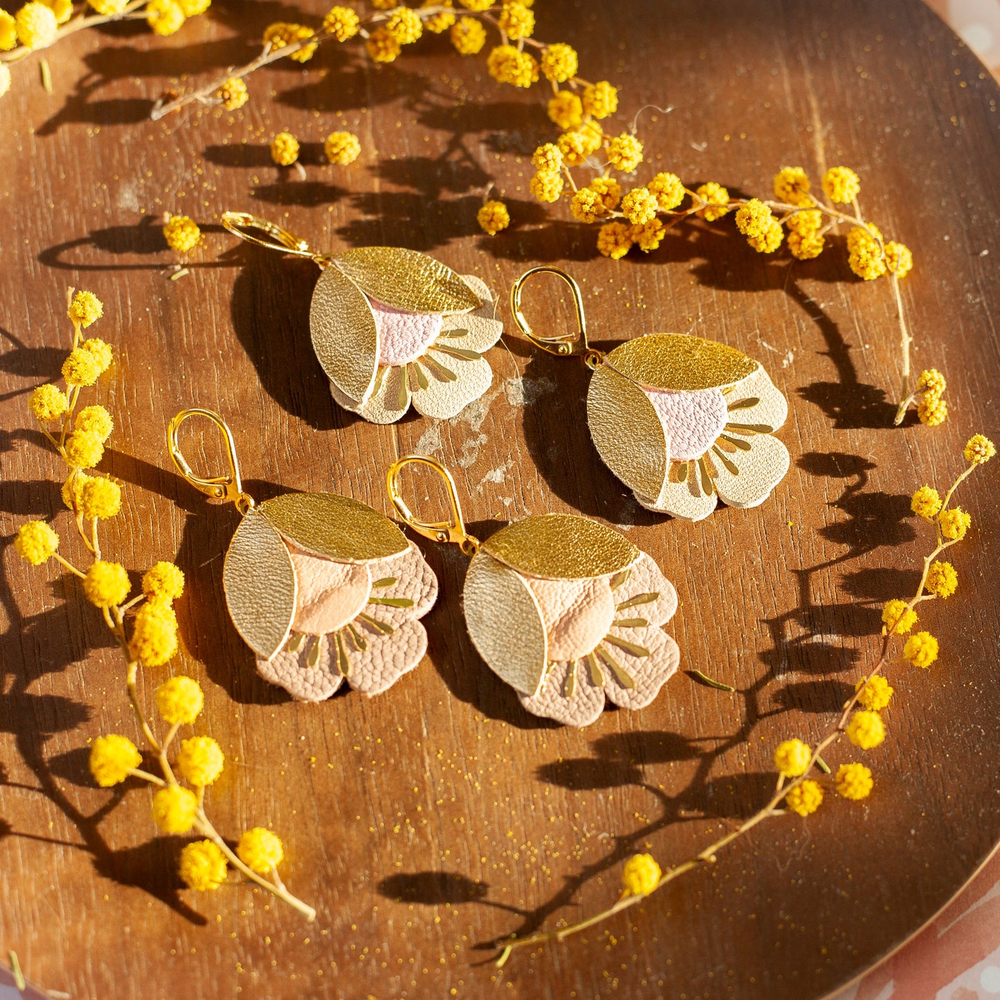 Boucles d'oreilles fleurs de cerisier doré et rose élégantes posées sur fond en bois avec décor floral jaune